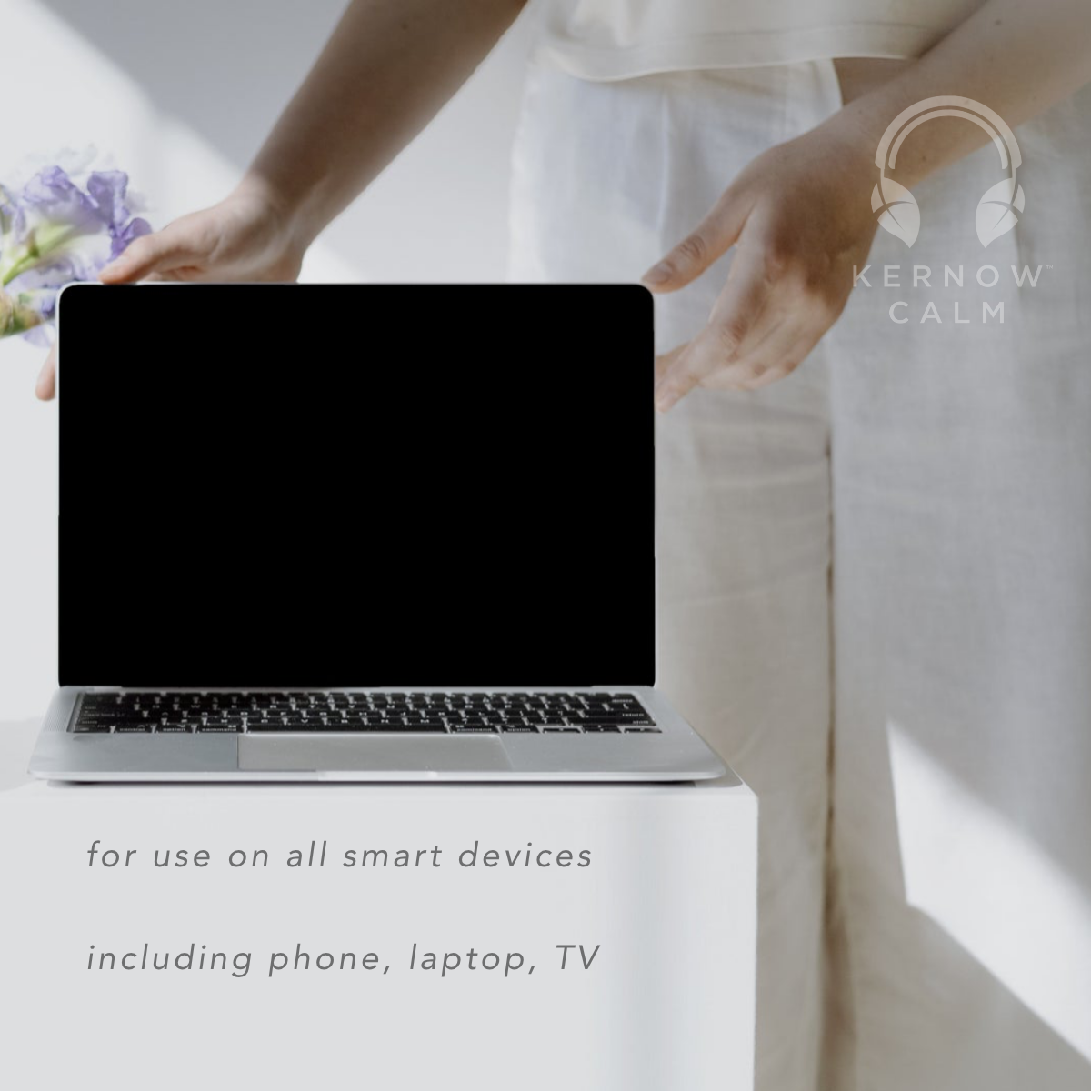 A silver laptop with a black screen shows compatible device list that sits on a white pedestal in a bright, minimalist setting. A person in white clothing is partially visible behind it, with hands reaching toward the laptop.